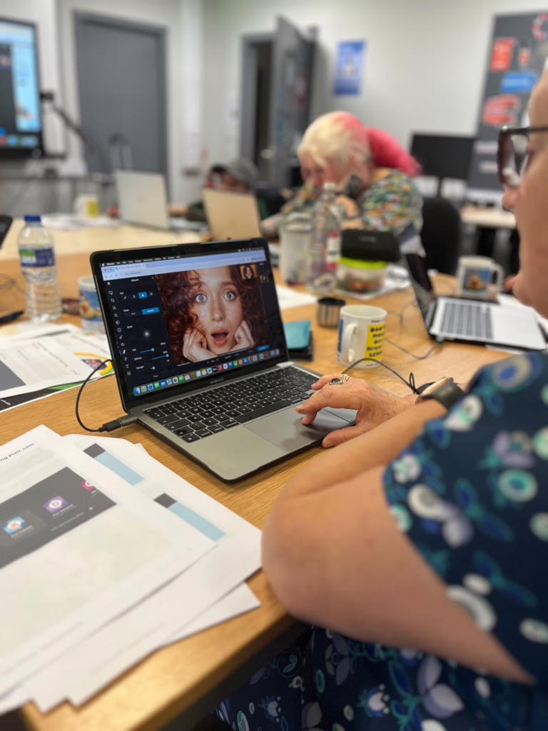 A person edits a photo using Pixlr during a creative workshop, surrounded by guides, mugs, and others working on laptops in a shared workspace.