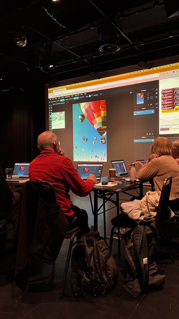 Participants follow along a photo editing demo using Pixlr, with hot air balloons on a large screen in a dark, theater-like creative workshop space.