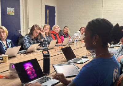 Participants sit around a long table with laptops, attentively joining a creative session led by a facilitator showing digital art on a screen.