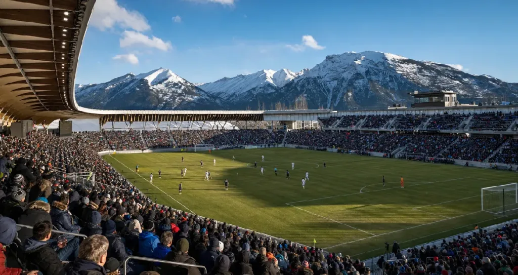 Football match in a mountain-side stadium with snow-capped peaks in the background, packed stands, and players on the pitch under clear daylight skies.