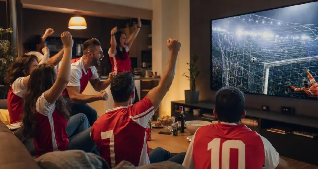 A group of football fans wearing matching red and white jerseys cheering excitedly in a cozy living room while watching a live soccer match on a large TV screen during a nighttime watch party.